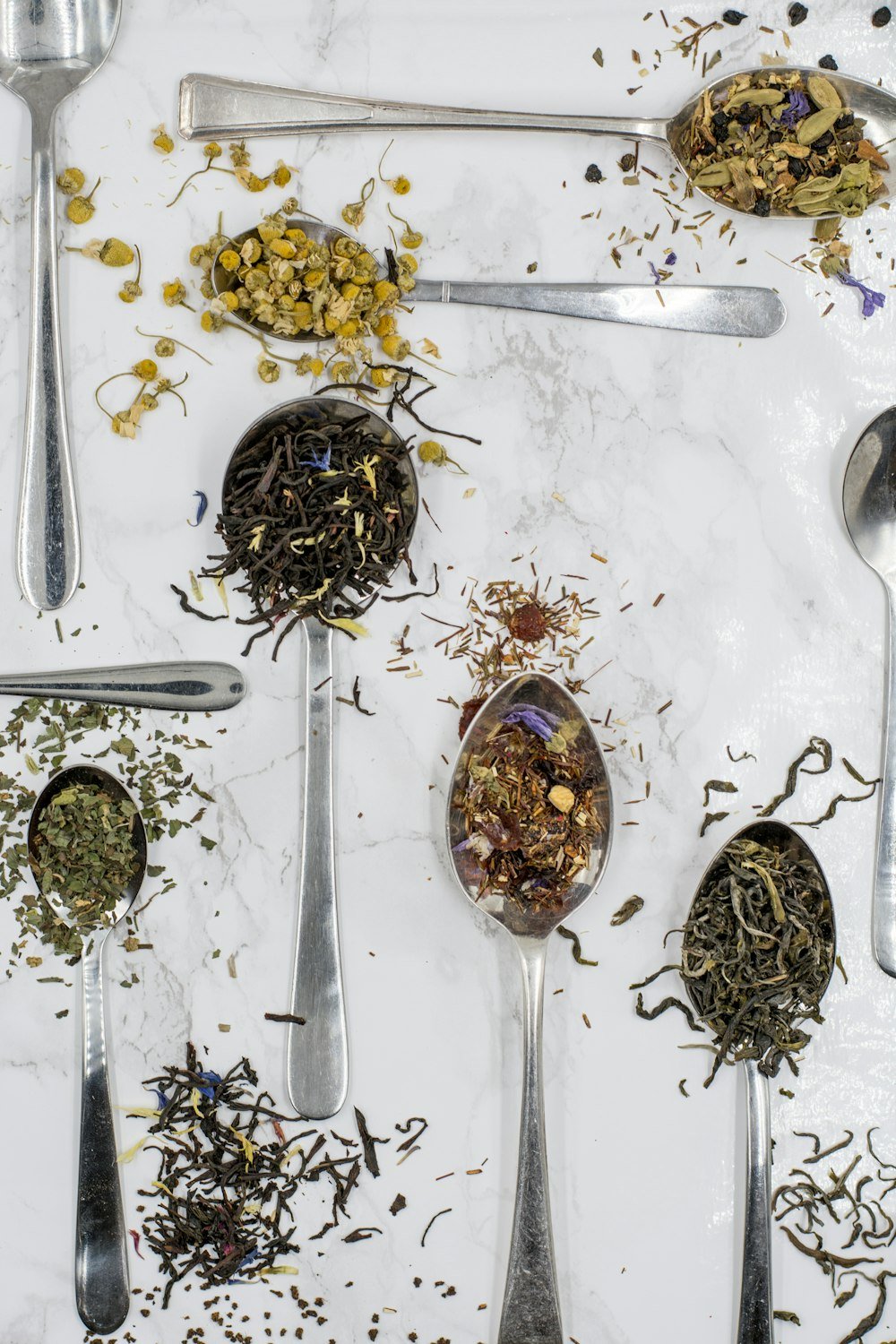 Spoons of dried herbs and loose-leaf teas on white marble